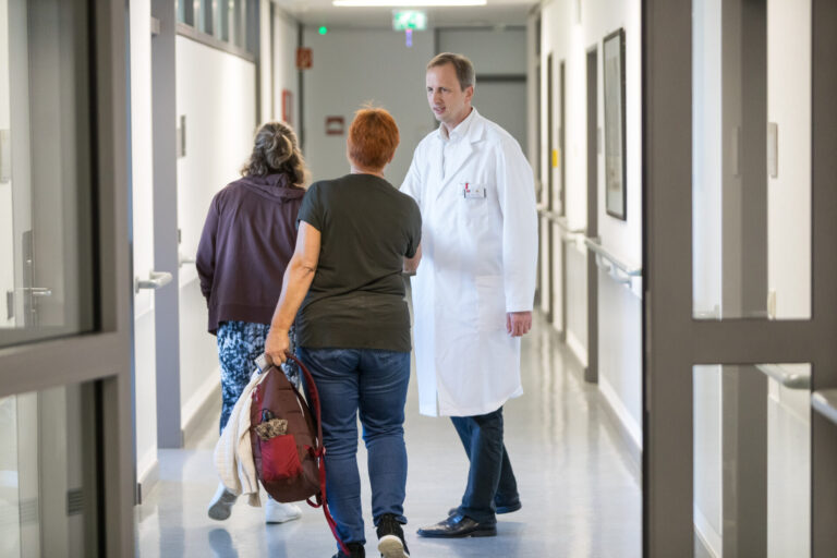 Arzt im weißen Kittel spricht mit zwei Patientinnen auf einem Krankenhausflur; eine Frau trägt eine Tasche, während die andere den Gang entlanggeht.