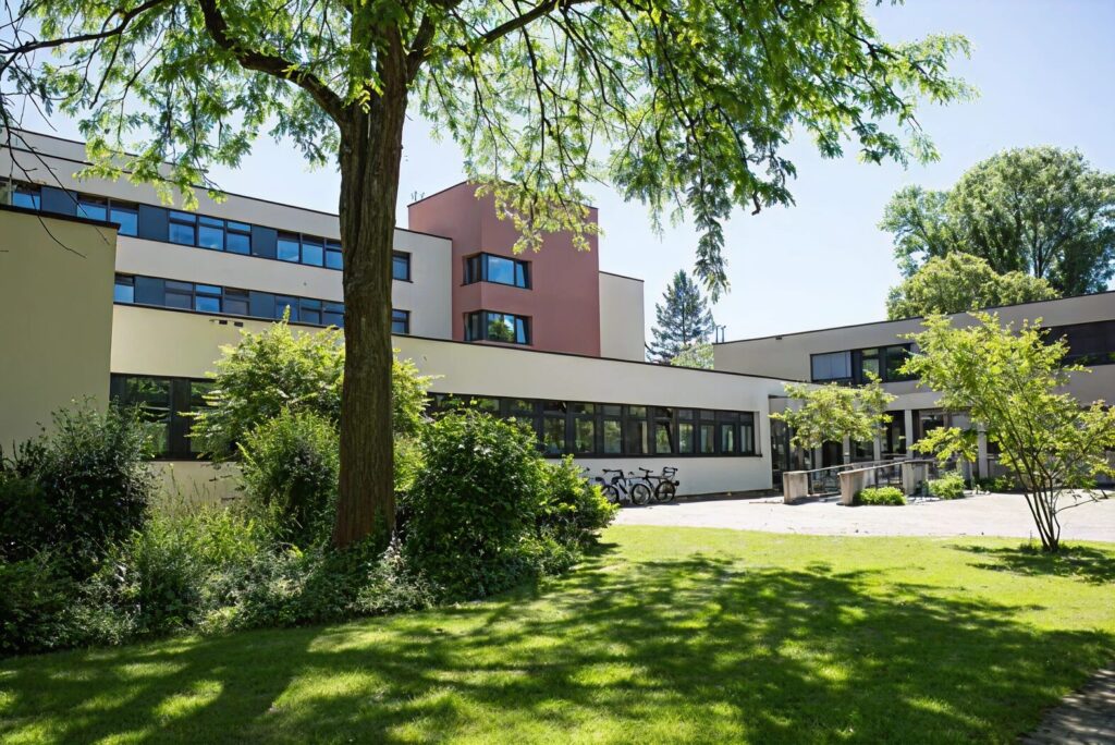 Modernes Klinikgebäude mit mehreren Flügeln und großen Fenstern, umgeben von Grünflächen, Bäumen und Fahrrädern im Innenhof bei sonnigem Wetter.