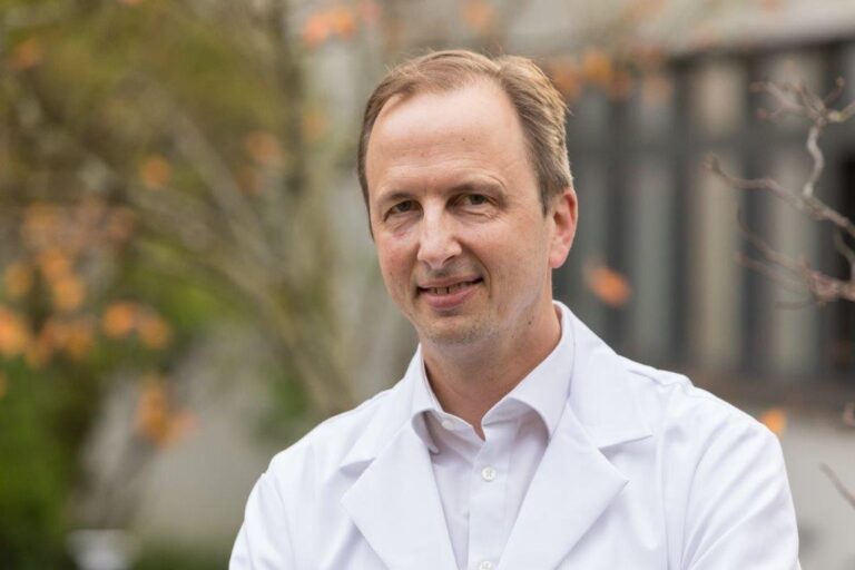 Porträt eines Arztes im weißen Kittel im Freien, vor unscharfem Hintergrund mit herbstlicher Bepflanzung.